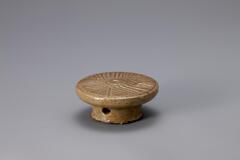 A small glazed porcelain piece resembling a cake stand. It has a wide top with a smaller and round vertical base. The top is detailed with a carved pattern that would imprint the rice cake being molded on it.<br />
<br />
Rice cake stamps are used to impress designs upon rice cakes. They are generally made of wood or ceramic. Ceramic rice cake stamps normally come in the form of round stamps and consist of a patterned surface and a handle. Patterns, carved or raised, on the stamp vary from geometric lines to auspicious designs that wish for prosperity and longevity. Their small size makes them highly portable, while their simple yet contemporary designs have mad them popular among collectors. The University of Michigan Museum of Art collection includes nine white porcelain rice cake stamps. Some are gifts from Mr. and Mrs. Hasenkamp, and others are gifts from Ok Ja Chang and the Chang family.<br />
<br />
This stamp is made of colored clay, which turned red after firing. It was thinly coated with a layer 