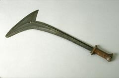 Metal blade with wooden handle. The metal blade is long and narrow and triangular at its end point. The wooden handle is split down the middle. Only one side of the blade has been decorated with long incisions. There is a small hole in the center of the triangular section of the blade. 
