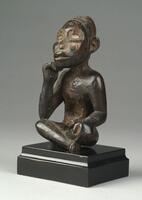 This figure sits serenely in a cross-legged position with one hand raised to the chin and the other resting upon on a knee. The figure's prominent rounded cheekbones, eyes embedded with pieces of mirror, large ears, and broad shoulders are typical traits seen in Vili carved figures. The figure is decorated with tukula powder and kaolin and has a worn, reddish-brown patina. The cavity at the back of the head and another in the abdomen indicate that this figure once bore potent medicinal substances and operated as an <em>nkisi</em>, an object of power.