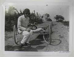 A photograph of a man resting in the middle of a dirt road. He sits on a homemade quadruped vehicle, made with bicycle wheels. He wears a riding cap and goggles.