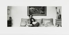 A black-and-white photograph of a couple on a couch. The man sits upright and the woman reclines, her head resting on his chest. A painting hangs behind them.