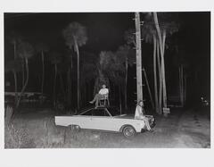 Photograph of a man in a folding chair on the roof and woman on the hood of a sedan at night in a forest setting.