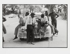 A black and white photograh of an old car, head on, with six men of varied age positioned on and around it. Houses and trees are along the street in the background. An elbow juts out of the left side of the frame.
