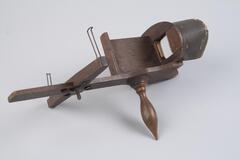 This stereoscope viewer is made of light brown wood and brass. It has an oval shaped view finder with two glass squares for the viewing holes. There is a handle below and a sliding wood arm with brass brackets to hold the stereoscopic card.