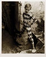 This portrait shows a young boy in a harlequin costume, looking into the camera lens. He stands in the ruins of Arles, arms akimbo, balancing on the ruins of the foundation of a building. 