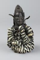 Figure of a human head on a base covered with cowrie shells. The cowrie shells are covered in blue pigment and two strings of cowrie shells hang off the back and side of the figure. There is leather wrapped around the neck of the figure. The head of the figure, carved out of wood, has pointed ears and a conical hairstyle decorated with geometric patterns and lines. 