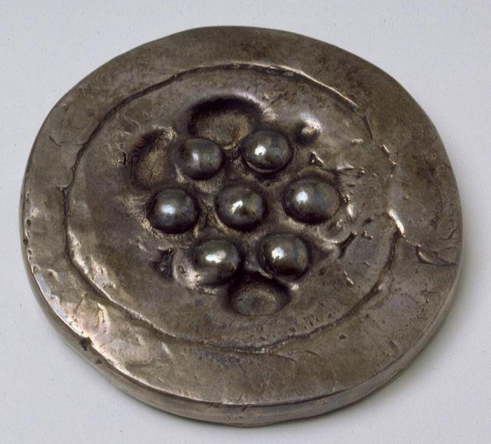 A small, thin silver disk. In the center are seven convex circular protrusions (one in the center surrounded symmetrically by the six others) that appear like balls set within individual depressions. There are other less uniform depressions surrounding the center design. An incised line runs around the circumference.