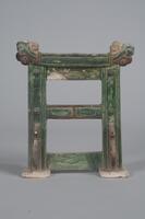 An earthenware miniature standing screen, mimicking wooden construction, with two vertical support beams and three cross beams on a platform with support brackets at the base. &nbsp;There are finials at the top on both sides, and it is glazed in green and amber.