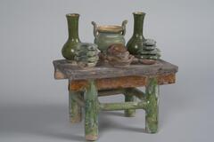 This is an earthenware miniature table glazed in brown and amber with green legs. Six objects are placed on the table in two rows of three. In the back row is a central <em>ding </em>censer with a green glaze; it is flanked by two two vases with globular bodies and tall straight necks. The front row has a central amber glazed bowl filled with a pig-head. This is flanked by green glazed bowls stacked with flat-shaped, possibly vegetal, offerings. 