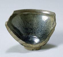 A fragment of a Jian Ware bowl&nbsp;with mottled black-brown glaze. It is on a footing and is dark on the bottom of the bowl and fades in to a lighter green around the rim.