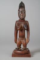 Standing female figure on a round base mounted on another square base. There are metal rings around the ankles. The hands are placed at the sides and there are strings of beads around the waist and neck. There are three vertical grooves in the forehead and on each cheek there are three vertical and three horizontal grooves. The hair is in a conical shape with a small knob at the top, decorated with vertical grooves.&nbsp;