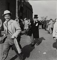 Image of people walking outside of a brick stadium. The man in the center of the image wears a big black coat and a top hat.