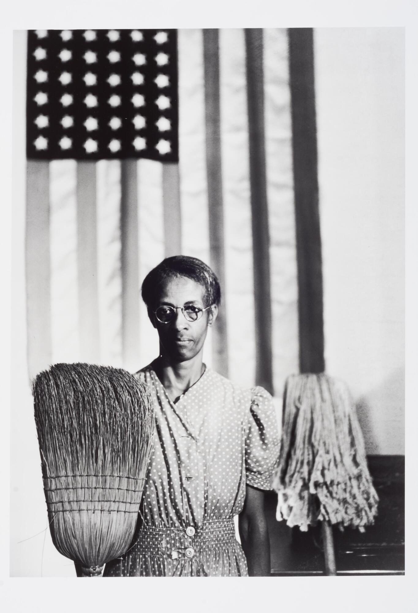 A person wearing a buttoned, polka-dot dress. They are standing in front of an American flag and behind a mop and broom.