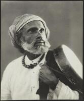 A black and white image of a bearded man in white garb, a tambourine draped over his left shoulder. &nbsp;He looks intensely upward and to the right.