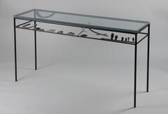 A metal table with a skeleton frame and a glass top. The underside of the table top has metal decorations of multiple shapes and figures extending toward the middle.