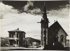A black and white photograph of a house on the left and church on the right, with numerous wires converging on the church&#39;s steep. Hills and mountains appear in the background.