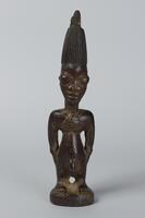 Standing male figure on a round base with both arms placed at the sides. There is a triangular carving, possibly an amulet, around the neck and back. The figure has a conical coiffure with vertical grooves. The coiffure terminates in two smaller conical projections. 