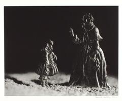 A photograph of two tin figures in a shallow plane, set against a dark, black backdrop. The small figure on the left stands facing the large figure on the right, as the figure on the left points her figure authoritatively.
