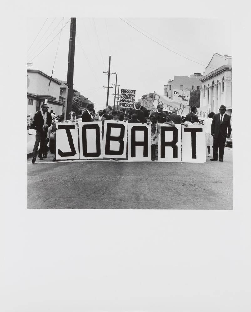 Black and white image of a group of people standing in a street and holding boards with letters to spell 'JOBART.'