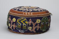 Circular cushion with multi-colored beadwork. The top is decorated with a large yellow flower surrounded by smaller flowers and vines on a blue background. The edge is outlined with red beads and the date &quot;1946&quot;. The sides are decorated with six alternating faces and yellow and red flowers. Three faces are green and red, while three are blue and yellow.