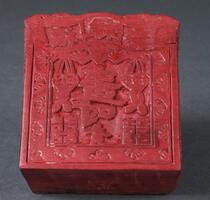 Wooden block painted cinnabar red with a carved floral design framing an inner square that contains characters in the top and lower right, center, and left corners and a large character in the middle with an image of two coins suspended from the wings of a stylized bat in the upper center.