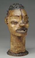Wooden mask in the shape of human head, covered with animal skin, possibly antelope. The head is ovoid shaped with a small nose and an open mouth filled with teeth. The ears are small and there is a dark line running from the top of the forehead to the bottom of the nose. There are two raised marks on each side of the face. The top of the head is darkened and has small, wooden pegs to represent hair. 