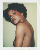 In this bust-length portrait, a woman poses nude, her body facing the left side of the frame, tilting her head toward toward the camera with her chin resting just above shoulder-level.