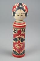A wooden doll with two tiers made up of a head and body with no arms, legs or feet. Painted on the head is a face, hair, and a red headdress. The body has red and black horizontal stripes on the top and bottom with a cluster of red and black abstract floral patterns.