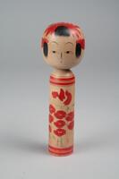 A wooden doll with two tiers made up of a head and body with no arms, legs or feet. Painted on the head is a face, hair, and a red headdress. The body has red and tan horizontal stripes on the top and bottom of the body with a cluster of red poppies in the middle.