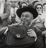 Image of a man wearing a wide-rimmed hat sitting amongst a crowd. He holds a bottle to his mouth and a satchel in front of him blocking most of the bottle.