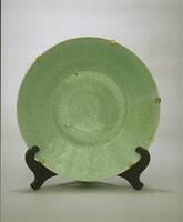 A stoneware flat bottomed plate on a footring with a wide flaring sides and direct rim.  The base is carved with a chrysanthemum and the sides with a peony meander.  It is covered in a green celadon glaze. This plate is a pair with 2002/2.6.