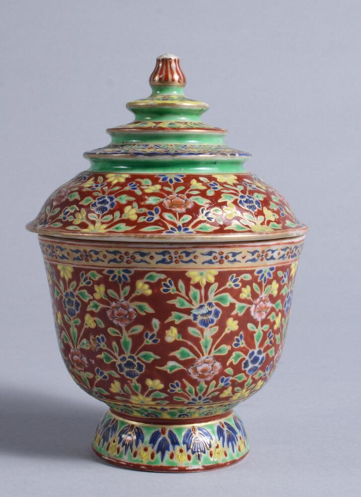 Covered ceramic jar with brilliantly colored Thai-inspired overglaze enamel painting in floral patterns.  The lid mimics the spires of Thai Buddhist architecture, rising from the gentle curve of the lower portion of the lid and alternating between solid bands of green, and multicolored floral patterns.  The green covers what would be the underside of each of the colorful three tiers of "roof" segments, culminating in what appears to be a red and gold lotus bud at the top of the lid.