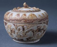 Small-size covered box, with a handle surrounded by a raised stylized calyx, which imitates the growth pattern of the top of a mangosteen, and a band of star points at the rim of the lid. The body has a band of vegetal scrolls. Brown-and-cream glaze.<br />