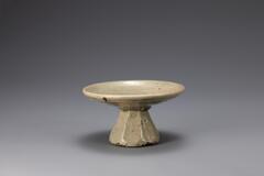 A glazed and speckled porcelain offering dish for an altar. The base is a wide decorated cylinder which tapers sharply into a narrow top. The base supports a wide, shallow bowl.<br />
<br />
This ritual dish was produced at a regional kiln. It is a low-quality object with a rough texture, made from the clay mixed with sand, contaminated with many impurities on the tray. Its glaze is dark with blue-green tints, giving the vessel the appearance of celadon. Its foot has an octagonal cross-section.<br />
[Korean Collection, University of Michigan Museum of Art (2014) p.200]