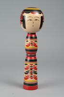 A wooden doll of three tiers made up of a head and body with no arms, legs or feet. The top of the doll is the head with a face, hair, and headdress of yellow and different shades of red painted on it. The second and third tiers make up the body and have yellow, black, and red decorations for clothing.
