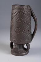 Vertically positioned cup with four loops at the base. The elongated cup handle has a face included. The cylindrical portion is carved with geometric intersecting lines forming a diamond pattern.