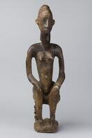 Female figure seated on a stool carved from a single piece of wood. The figure has horizontal marks on the chest and upper arms. The figure&#39;s head is long and thin with protruding features and carved &#39;hair&#39; extending over the head and down the back of the neck. The figure&#39;s left arm has remnants of a cuff made of animal hide. The stool is four-legged and the back legs are carved figures.&nbsp;
