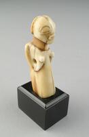 As one of a pair of delicately carved, ivory statuette-pendants, this female figure stands upright, as opposed to her counterpart whose head and upper body lean slightly forward. Both, however, have a round head with a convex face; large, coffeebean-shaped eyes; a rectangular mouth with prominent lips; a cylindrical neck; and, a coiffure decorated on the back with a cruciform pattern. Additionally, both female figures clutch their breasts in their hands. The statuettes have been pierced through, allowing them to suspend from a string.