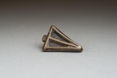 Gold-weight in the shape of a triangular base, with a raised line along the edges and a bisecting line down the middle. Along one edge of the base there is a small circular protrusion. 