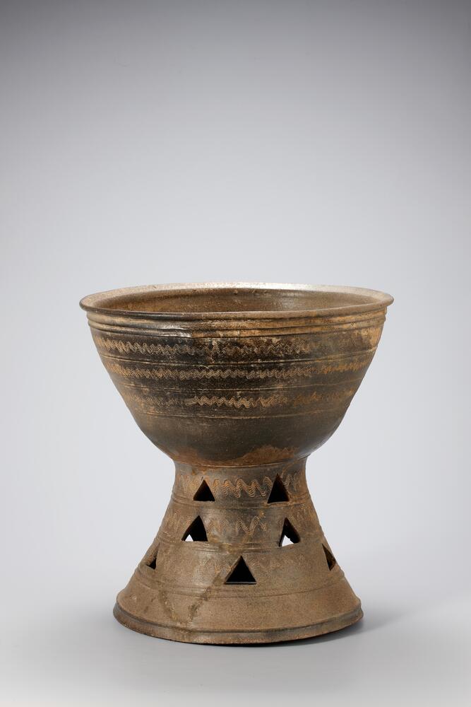 Large pedestal bowl with flaring mouth and base cut with evenly spaced triangular designs. Three side-by-side horizontal bands decorate the lip of the bowl, while six bands are spaced along the sides encompass the exterior of the bowl. In between these bands incised wavy lines stretch around the body horizontally.<br />
<br />
This is a well-fired, dark gray, bowl-shaped vessel stand. A set of two thick raised bands encircles the stand just below the rim, and three horizontal ridges divide the surface of the bowl below the bands. The sections divided by these bands and ridges have been decorated with wave designs rendered using a four-tooth comb. The bowl has an everted mouth and a round rim. The pedestal is also divided by raised bands into sections decorated with wave designs. Each of the upper two sections features five triangular perforations, while the lower sections feature five triangular perforations alternately offset from those of the sections above. The inner and outer surfaces show faint traces 