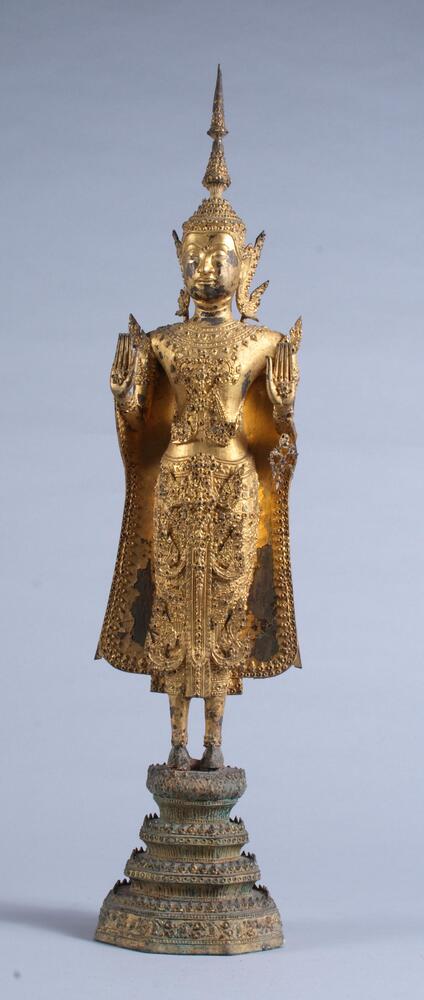 Standing gilt bronze Buddha with glass inlay in royal costume.  Stands on a lotus pedestal with both hands raised in abhaya mudra.