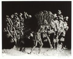 A photograph of a tin figure in a shallow plane, set against a black backdrop. Large plants surround the male figure. He holds a large basket on his shoulder, above his head, carrying it and leaning forward, illustrating the burden of the large load.