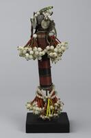Wooden columnar figure wrapped in alternating black and red wire. The top of the figure is wrapped in wire and decorated with leather strands of white beads and a 1946 coin. The limbs are formed by strands of green and red seed beads terminating in large white beads.  