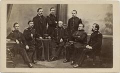 A photograph of eight men in military garb, five seated and three standing, arranged in a shallow arc in an interior studio setting. 