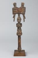 Staff with a cylindrical handle and a kneeling female figure on a small, disc-shaped base. The figure is holding her breasts in both hands and her hair is decorated with vertical grooves. The top of the figure&#39;s head is surmounted by a double-axe shaped form, decorated with six incised lozenges. At the top of the double-axe form are two rectangular protrusions and at the bottom are two protrusions that may represent lightning bolts.&nbsp;