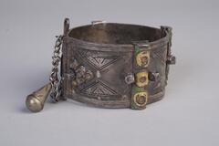 A silver cuff bracelet with braided carvings and green and yellow decorations.  A chain is attached to one side to keep it closed.