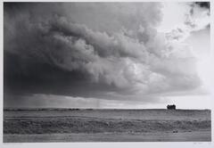 A photograph of a large, dark stormcloud and tornado rolling over a flat plain. A barn is visible on the horizon line.