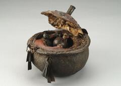 This vessel is formed from a hollowed calabash or gourd, the top of which has been carved to form a lid, decoratively patterned with brass tacks and studded by a handle at the center. Snake skin covers the rounded base of the calabash conferring a coarse texture. Several holes have been drilled around the top perimeter of the base from which metal rings holding iron clappers or bells dangle. An array of man-made, animal, and vegetal objects are held inside the vessel, namely, small, wooden anthropomorphic figurines carved with a minimum of physiognomic detail, bird claws, feathers, wings, split cane, and dried seeds.&nbsp;
