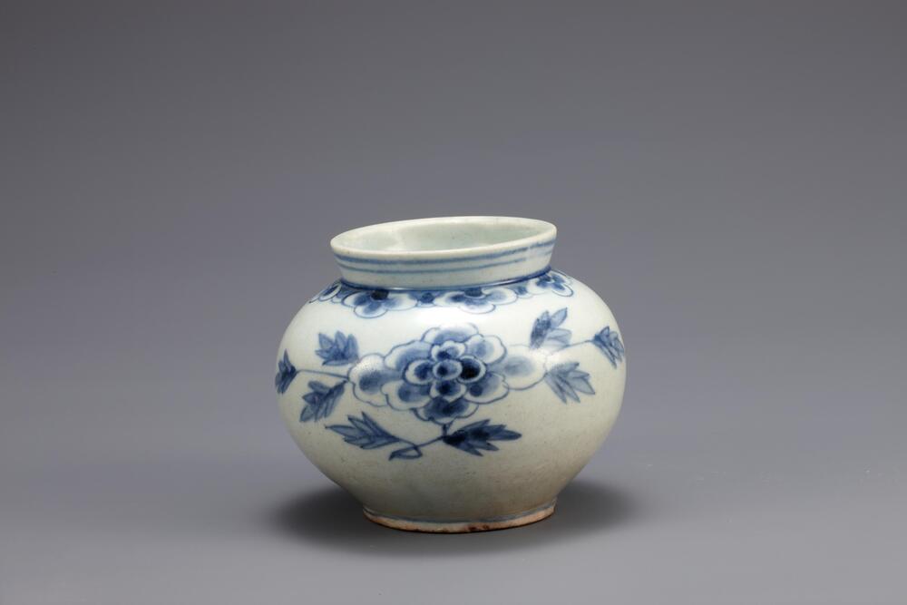 This small jar features large peony sprays in the center of the body, and above them are decorated a yeoui-head band. This type of the small blue-and-white porcelain jars was popular among collectors thanks to the outstanding harmony of white and blue. This piece has coarse sand spur marks, and red spots are visible on the foot rim. The jar remains intact, and the glaze was well fused.<br />
[Korean Collection, University of Michigan Museum of Art (2014) p.173]
