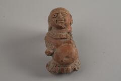 An Indian terracotta figure with big eyes and distinguishable facial features. The figure has a big tummy and the left arm of the figure is borken and gone.
